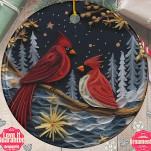 May include: A round ceramic ornament featuring a quilling-style illustration of two red cardinals perched on a branch against a dark blue winter forest background with white snow, pale trees, and yellow star-shaped accents.