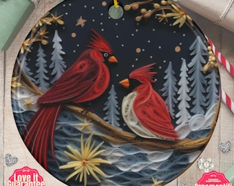 May include: A round ceramic ornament featuring a quilling-style illustration of two red cardinals perched on a branch against a dark blue winter forest background with white snow, pale trees, and yellow star-shaped accents.
