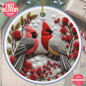 May include: A round ceramic ornament featuring two cardinals perched on a branch with red berries. The birds have red and gray feathers, with black beaks. The ornament has a blue border and a gold string for hanging. The text "Fast Delivery" and "Ornmentifi" are visible.