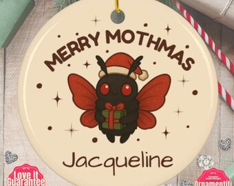 May include: A round ceramic ornament with a cream background featuring a black moth character wearing a red Santa hat and holding a green gift box. The text MERRY MOTHMAS is printed above the moth, and the name Jacqueline is printed below.