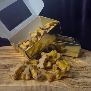Op de afbeelding: Een gouden doos gevuld met hondenkoekjes in de vorm van botten. De doos is open en de koekjes vallen op een houten oppervlak.