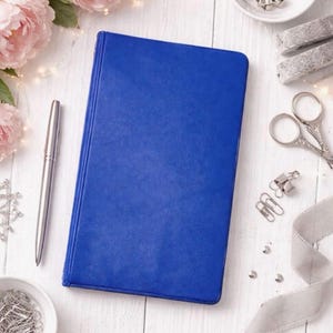 May include: A cobalt blue journal lies on a white wooden surface, next to a silver pen, pink flowers, and stationery supplies. The journal is closed and has a smooth cover. Other items include paper clips, scissors, and decorative ribbon.