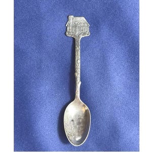 May include: Silver-toned spoon with a detailed house design at the top. The handle has a textured, branch-like appearance, and the spoon bowl is oval. The spoon is set against a blue background.