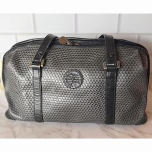 May include: A gray travel bag with a geometric pattern and black leather straps. The bag has a top zipper closure and a circular emblem on the front. The bag measures approximately 12 inches long and 8 inches high.