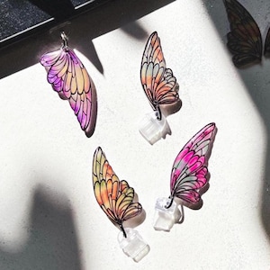Può includere: Quattro orecchini a forma di ali di farfalla colorate nelle tonalità del rosa, viola, arancione e giallo. Le ali sono dettagliate con contorni neri e sono attaccate a perni trasparenti per orecchini. Sullo sfondo è visibile un paio di ali più scure.