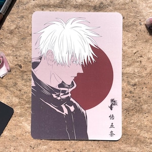May include: A rectangular card featuring an anime-style illustration of a character with white hair and a dark outfit. The background includes a red circle and Japanese text. The artwork is in shades of gray, black, and red.