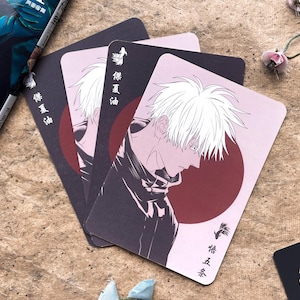 May include: A stack of anime-style playing cards featuring a character with white hair and a dark outfit against a pink and burgundy background. Japanese text is visible on the cards. A book and other decorative items are in the background.
