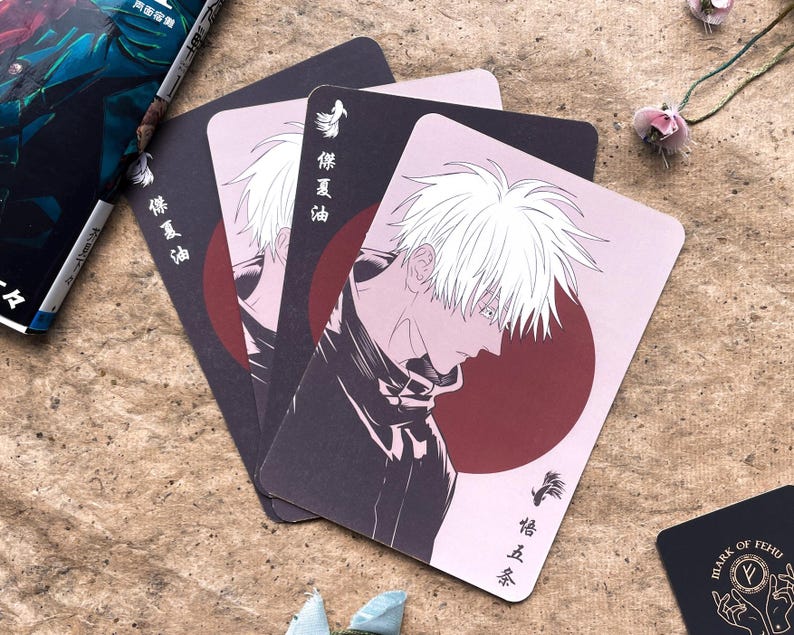 May include: A stack of anime-style playing cards featuring a character with white hair and a dark outfit against a pink and burgundy background. Japanese text is visible on the cards. A book and other decorative items are in the background.
