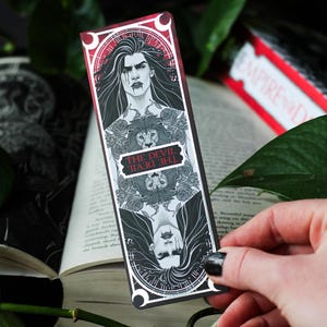 Puede incluir: Un marcapáginas en blanco y negro con una ilustración gótica de una persona y un león. El marcapáginas tiene el texto "THE DEVIL" en rojo y blanco. El marcapáginas se sostiene sobre un libro abierto con hojas verdes en el fondo.