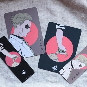 May include: Four illustrated cards and bookmarks featuring anime-style characters. The designs include a man in a white coat and a hand with a snake. The color palette includes black, pink, and gray.