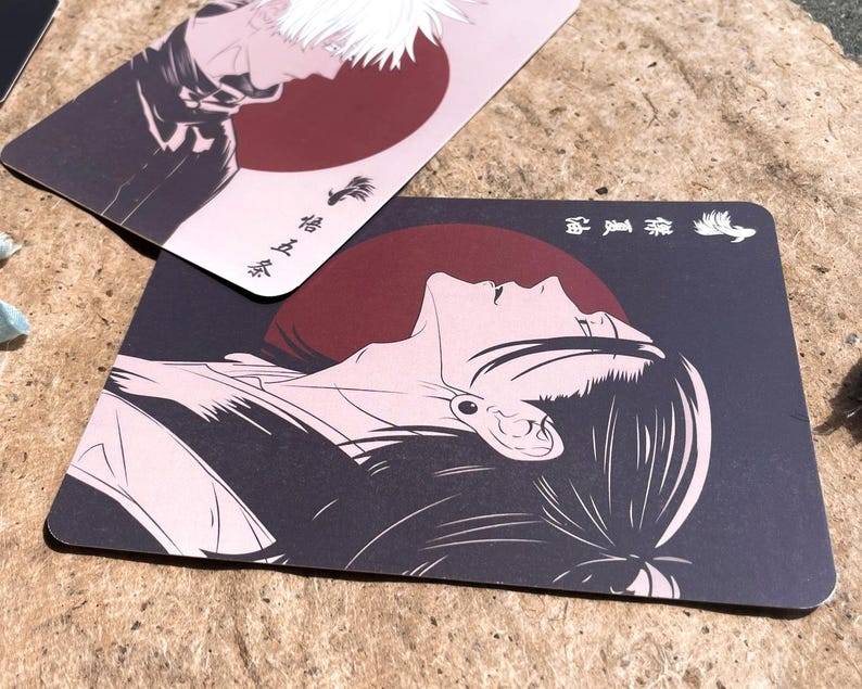 May include: Two rectangular cards with anime-style illustrations. One features a profile of a character with white hair against a red circle. The other card shows a person's face looking up, also with a red circle background. Japanese text is visible.