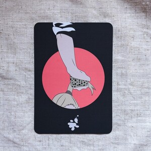 May include: A black rectangular card with an illustration of a hand, a red circle, and a figure. The hand is holding a dotted object. A white splash is at the bottom. The background is a light gray fabric.