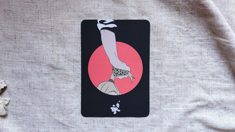 May include: A black rectangular card with an illustration of a hand, a red circle, and a figure. The hand is holding a dotted object. A white splash is at the bottom. The background is a light gray fabric.