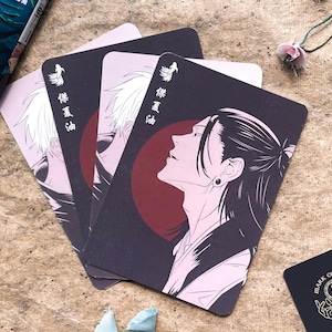 May include: Set of three playing cards featuring anime-style illustrations. Each card has a dark background with a character portrait, a red circle, and Japanese text. A small black card with white text is also present.