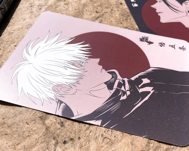 May include: A light pink card featuring a white-haired anime character in profile against a dark red sun. The character is wearing a dark jacket. The card has Japanese text and is set on a textured surface.