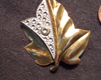 May include: A gold and silver leaf-shaped brooch with a cluster of rhinestones in the center.