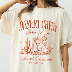 May include: Ivory-colored t-shirt with a red graphic that reads "DESERT CREW Before I Do" with a cactus, margarita, and perfume bottle design. The text "Jaclyn's Bachelorette Scottsdale, AZ" is also printed on the shirt. The shirt is a loose fit.