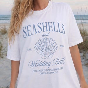 May include: White t-shirt with blue text and seashell graphic. The shirt reads "Seashells and Wedding Bells" with "Chelsea's Bachelorette Charleston, SC" below. The shirt is worn on a beach.