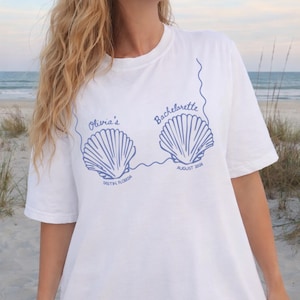 May include: White t-shirt with a blue seashell design. The shirt features the text "Olivia's Bachelorette" with the location "Destin, Florida" and the date "August 2024". The shirt is made of a soft material.
