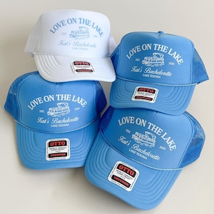 May include: Four trucker hats in white and light blue with mesh backs and foam fronts. Each hat features a graphic of a pontoon boat with the text LOVE ON THE LAKE, Kat's Bachelorette, and LAKE TEXOMA. Each hat has an Otto brand sticker on the brim.