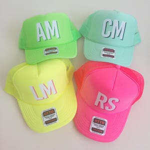 May include: Four trucker hats in neon green, mint green, neon yellow, and neon pink. Each hat features a foam front panel with white block letters: AM, CM, LM, and RS. Each hat has a mesh back, a curved brim, and an Otto brand sticker on the front.