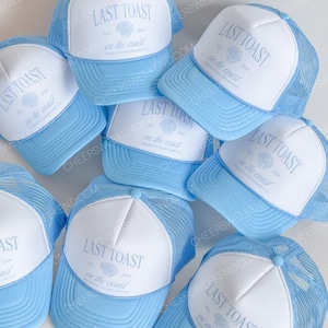 May include: A collection of light blue and white trucker hats. Each hat features a white front panel with the text "LAST TOAST" and "on the coast" printed in light blue, along with a seashell graphic. The hats have a mesh back.