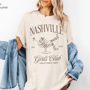 Pu&ograve; includere: Una t-shirt color avorio con una stampa grafica color espresso che riporta il testo Nashville, Girls Club, Est. 2025 e Ashley's Dirty Thirty. Il design include un'illustrazione a linee di una persona con cappello e stivali da cowboy dentro una coppa da martini.