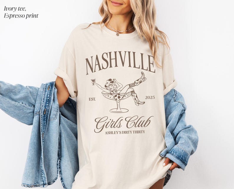 Pu&ograve; includere: Una t-shirt color avorio con una stampa grafica color espresso che riporta il testo Nashville, Girls Club, Est. 2025 e Ashley's Dirty Thirty. Il design include un'illustrazione a linee di una persona con cappello e stivali da cowboy dentro una coppa da martini.