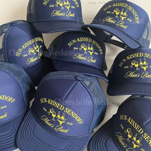 May include: Navy blue trucker hats with yellow text that reads "SUN-KISSED SENDOFF JUNE 2026 Maia's Bach". The hats have a mesh back and a curved brim. The hats are likely for a bachelorette party.
