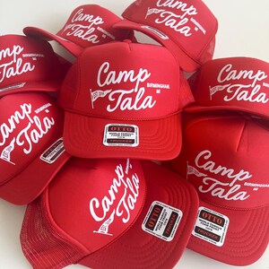 Puede incluir: Una colecci&oacute;n de gorras de camionero de color rojo brillante con un logotipo serigrafiado en blanco que dice Camp Tala Birmingham MI, un peque&ntilde;o gr&aacute;fico de bander&iacute;n y el texto EST 2016. Cada gorra tiene frente de espuma y etiqueta Otto.