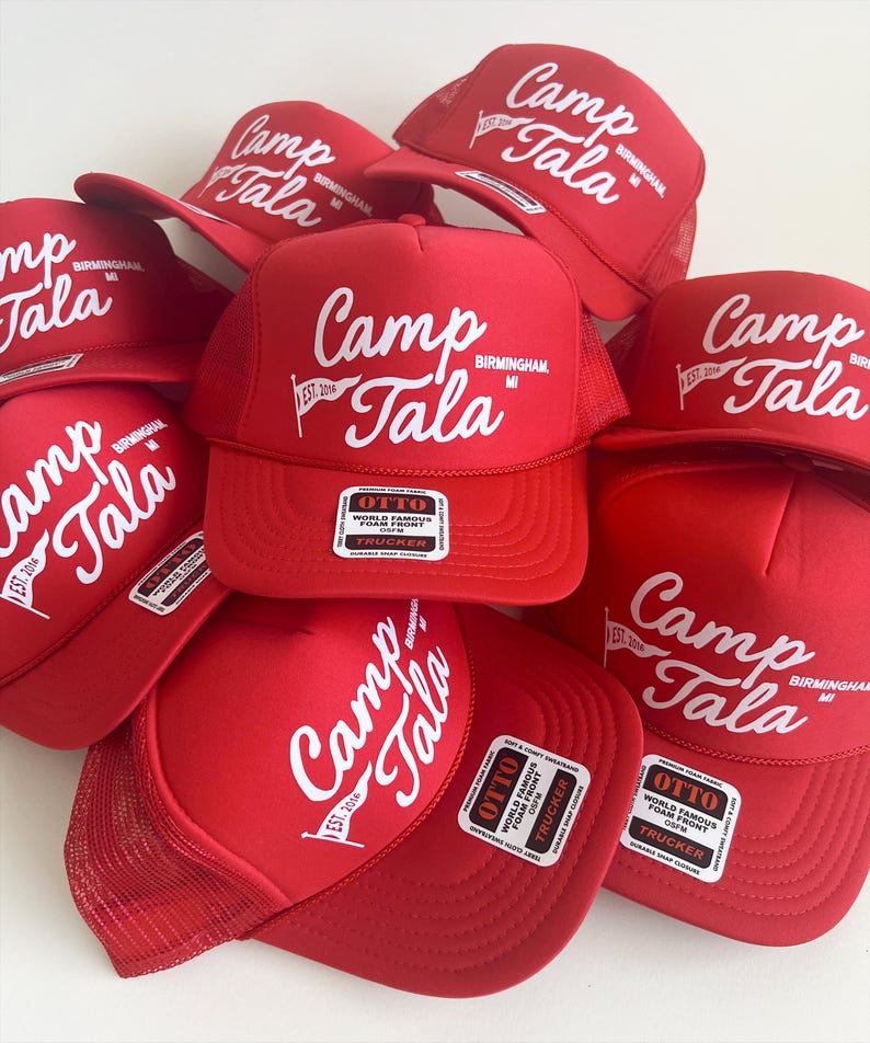Puede incluir: Una colecci&oacute;n de gorras de camionero de color rojo brillante con un logotipo serigrafiado en blanco que dice Camp Tala Birmingham MI, un peque&ntilde;o gr&aacute;fico de bander&iacute;n y el texto EST 2016. Cada gorra tiene frente de espuma y etiqueta Otto.