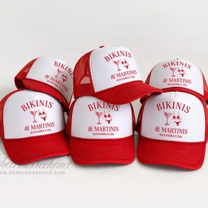 May include: A stack of red and white trucker hats. The hats have a red brim and mesh back, with a white front panel. The front panel of each hat displays the text "BIKINIS & MARTINIS" in red, along with a martini glass and bikini graphic.
