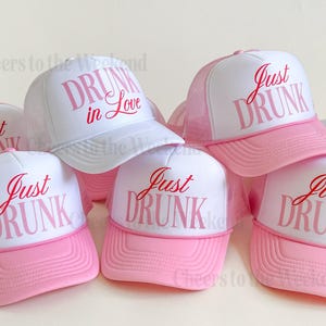May include: A collection of white and pink trucker hats. The hats have a white front panel with the words "Just DRUNK" or "DRUNK in Love" in pink script. The brim and mesh backing are pink.