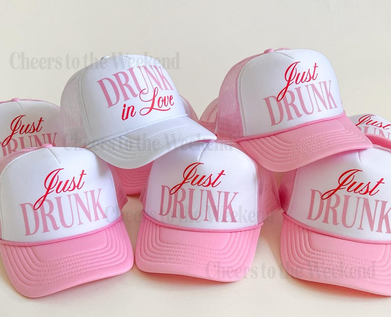 May include: A collection of white and pink trucker hats. The hats have a white front panel with the words "Just DRUNK" or "DRUNK in Love" in pink script. The brim and mesh backing are pink.