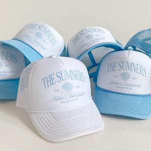 May include: White trucker hats with light blue brims and mesh backs. The hats feature the text "THE SUMNERS" in light blue, along with "Ashton's Bachelorette" and "OAK ISLAND, NC". A seashell graphic is also included.