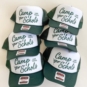 Puede incluir: Un conjunto de cinco gorras de camionero de color verde oscuro y blanco con un panel frontal de espuma blanca con el texto en verde oscuro Camp Echols Lake Travis y un icono de bandera con el texto EST. 2027. Cada gorra tiene una malla trasera.