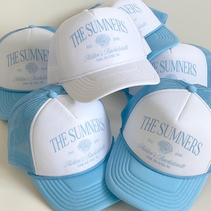 May include: A collection of white and light blue trucker hats featuring a printed design with the text THE SUMNERS, EST. 2025, a seashell graphic, and the script text Ashton's Bachelorette OAK ISLAND, NC. Each hat has a mesh back and a solid brim.