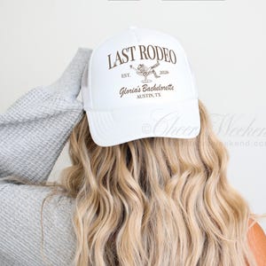 May include: White trucker hat with the text "LAST RODEO" in brown, along with a graphic of a person in a cowboy hat. The hat also includes the text "Gloria's Bachelorette, Austin, TX".