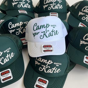 May include: A collection of trucker hats featuring a foam front and mesh back. Most hats are dark green with white text, while one is white with dark green text. Each hat displays the text Camp Katie, Seattle, WA, and a small pennant icon.