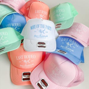 K&ouml;nnte beinhalten: Eine Sammlung von Trucker-Caps mit Schaumstofffront in Pastellrosa, Koralle, Mintgr&uuml;n und Blau. Die meisten Kappen zeigen den Schriftzug "LAST RODEO" mit einem Rodeo-Grafikmotiv, w&auml;hrend eine wei&szlig;e Kappe den Text "WIFE OF THE PARTY" in Blau tr&auml;gt.