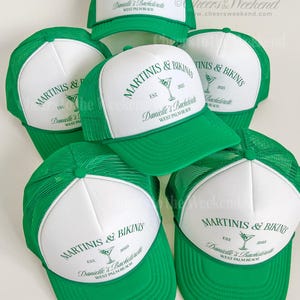 May include: A collection of green and white trucker hats. Each hat features the text "MARTINIS & BIKINIS" and "Danielle's Bachelorette WEST PALM BEACH" in green. The hats have a mesh back and a curved brim.