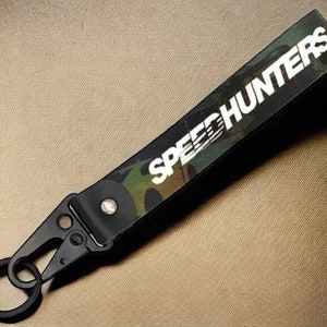 Puede incluir: Llavero de camuflaje negro y verde con un clip de metal negro y un logotipo "SPEED HUNTERS" blanco.