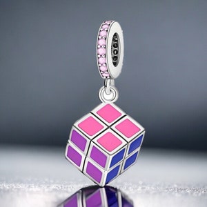 May include: A silver charm in the shape of a Rubik's Cube with pink, purple, and blue enamel. The charm has a silver ring with pink stones around it.