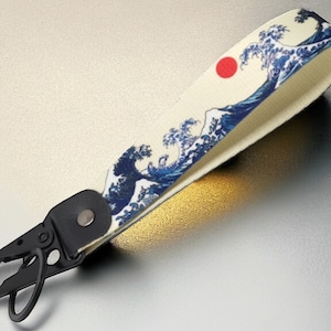 Può includere: Un portachiavi bianco e nero con un design a onde blu e bianche e un cerchio rosso. Il design ricorda la Grande Onda di Kanagawa.