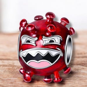 Puede incluir: Un charm rojo con un diseño de virus de dibujos animados. El charm tiene un cuerpo redondo con púas que sobresalen y una cara con una amplia sonrisa dentuda. Los detalles plateados delinean la cara y los bordes del charm.