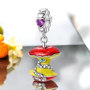 Könnte beinhalten: Ein silberner Anhänger in Form eines angebissenen Apfels mit roter und gelber Emaille. Der Apfel hat eine grüne Schlange, die sich um ihn herumwickelt. Der Anhänger hat ein silbernes Herz mit einem violetten Stein oben.