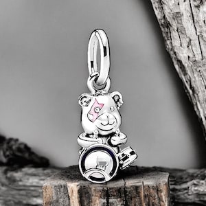 May include: A silver charm featuring a cartoon bear playing the drums. The bear has a pink star on its eye and is wearing a blue and white drum set.