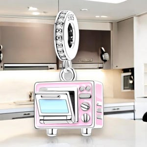 May include: A silver charm in the shape of a pink microwave with a silver door and silver details. The charm is attached to a silver chain with a circle of clear crystals.