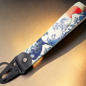 Könnte beinhalten: Ein schwarzer Schlüsselanhänger aus Metall mit einem weißen und blauen Stoffband mit einem japanischen Wellendesign. Das Band hat eine rote Sonne, die über der Welle aufgeht.