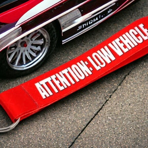 Puede incluir: Llavero rojo con un anillo blanco y el texto "ATENCIÓN: VEHÍCULO BAJO" en letras blancas.